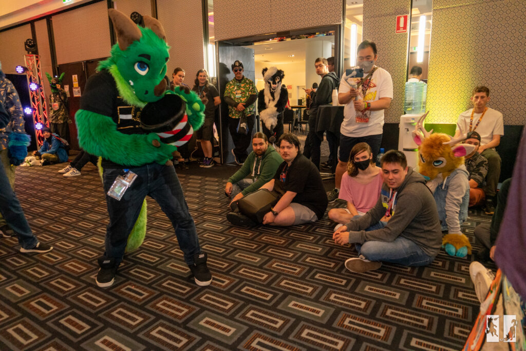 a green fursuiter playing a game with a bucket surrounded by onlookers
